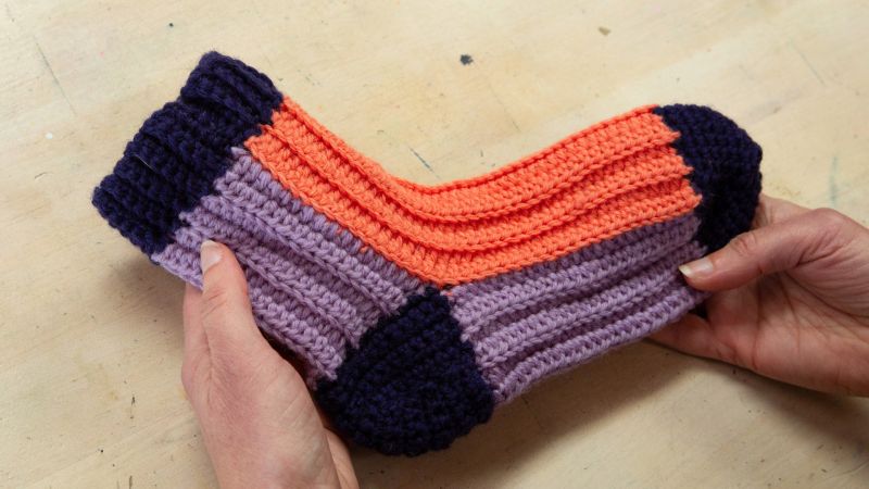 Two hands hold a knitted slipper sock with purple, orange, and dark blue stripes on a light wooden surface.
