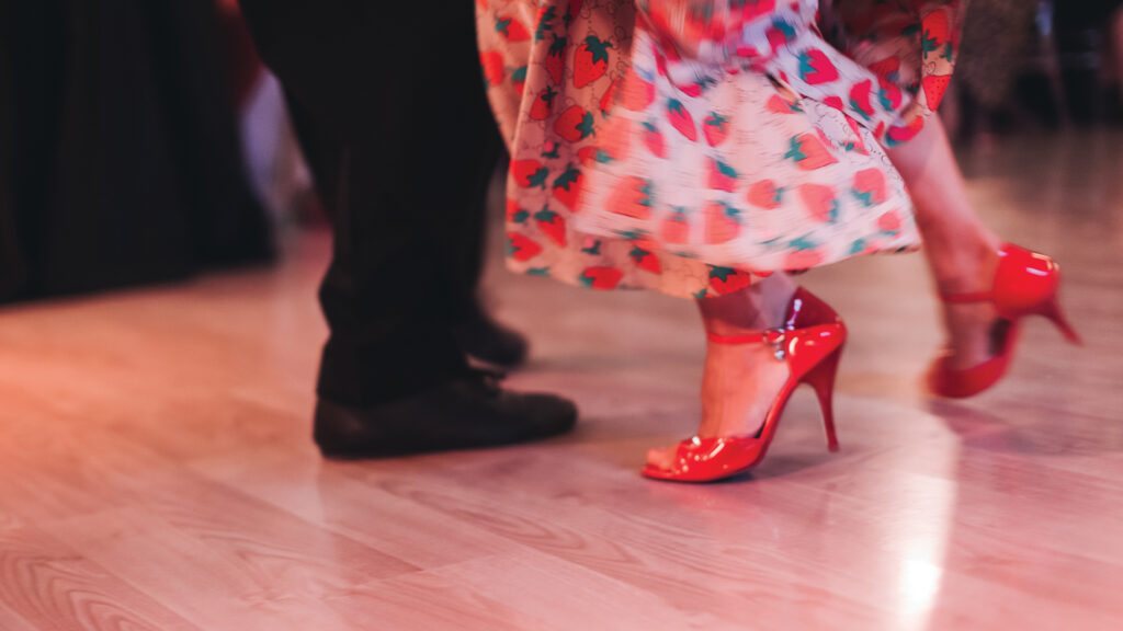 Two people dancing; one in red high heels and a colorful dress, the other in dark pants and shoes.