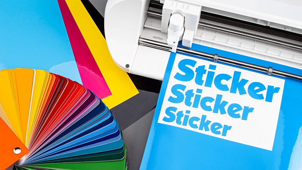 A vinyl cutter with sheets of colored paper and a printed sheet saying "Sticker" multiple times.