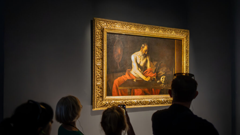 People view and photograph a framed painting of a man writing at a table in a dimly lit gallery.