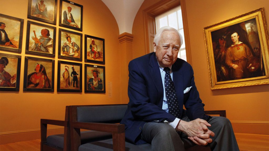 A man in a suit sits on a bench in an art gallery, with several portraits on the wall behind him.