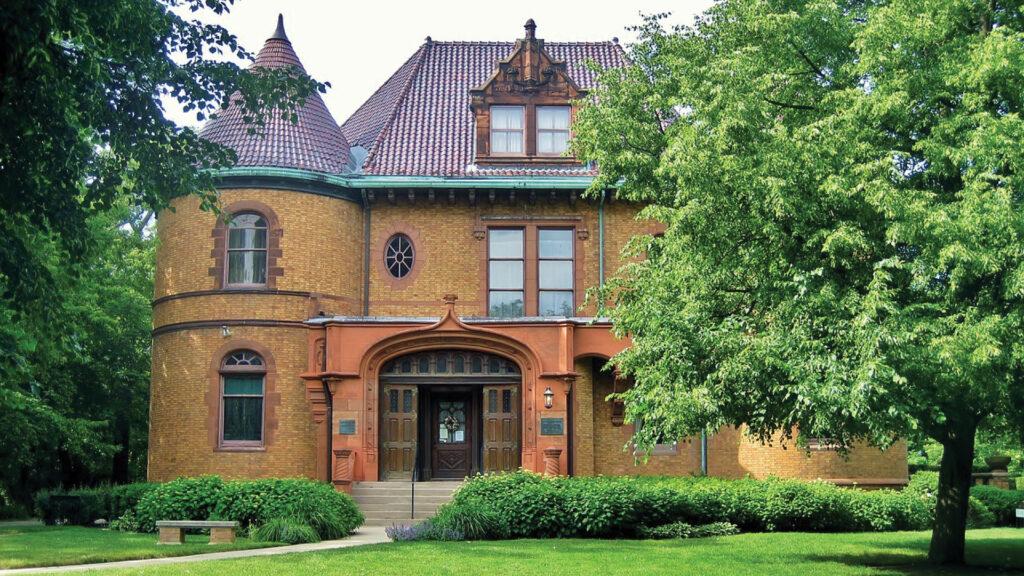 Historic brick mansion with a steep roof and round turret, surrounded by lush greenery.