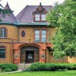 Historic brick mansion with a steep roof and round turret, surrounded by lush greenery.