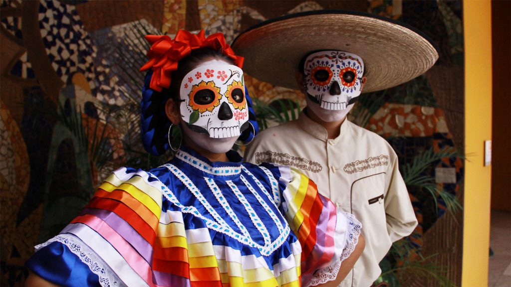 Two people dressed in traditional Day of the Dead costumes with colorful face paint and attire standing in front of a mural.