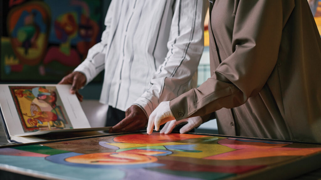 Two people examine colorful abstract artwork on a table, one wearing white gloves and turning a page.