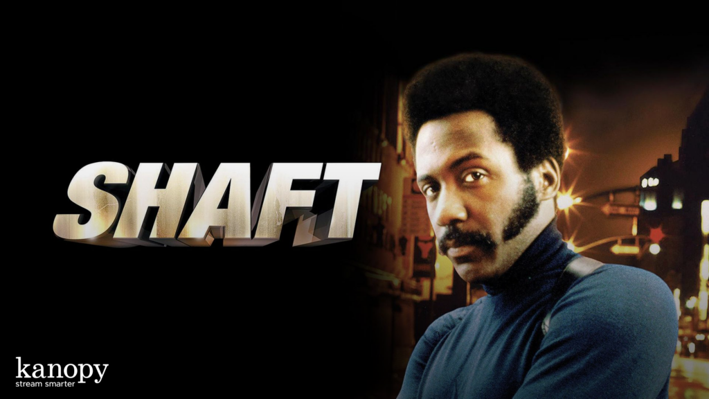 Man in blue turtleneck stands next to large “SHAFT” text with city lights in the background.