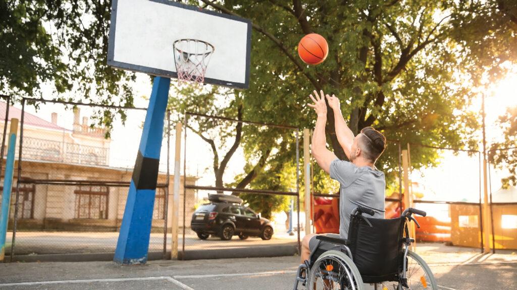 Man in a wheelchair shooting a basketball towards a hoop on an outdoor court.