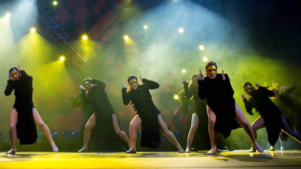 Group of dancers in black costumes striking dramatic poses on a foggy, lit-up stage.