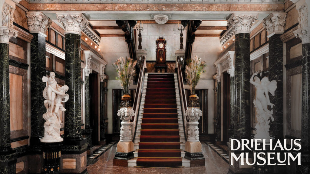 Grand marble staircase with ornate columns, statues, and the Driehaus Museum logo in the bottom right corner.