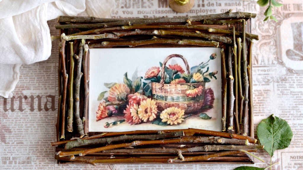 A twig-framed painting of a basket filled with orange and yellow flowers on a newspaper background.