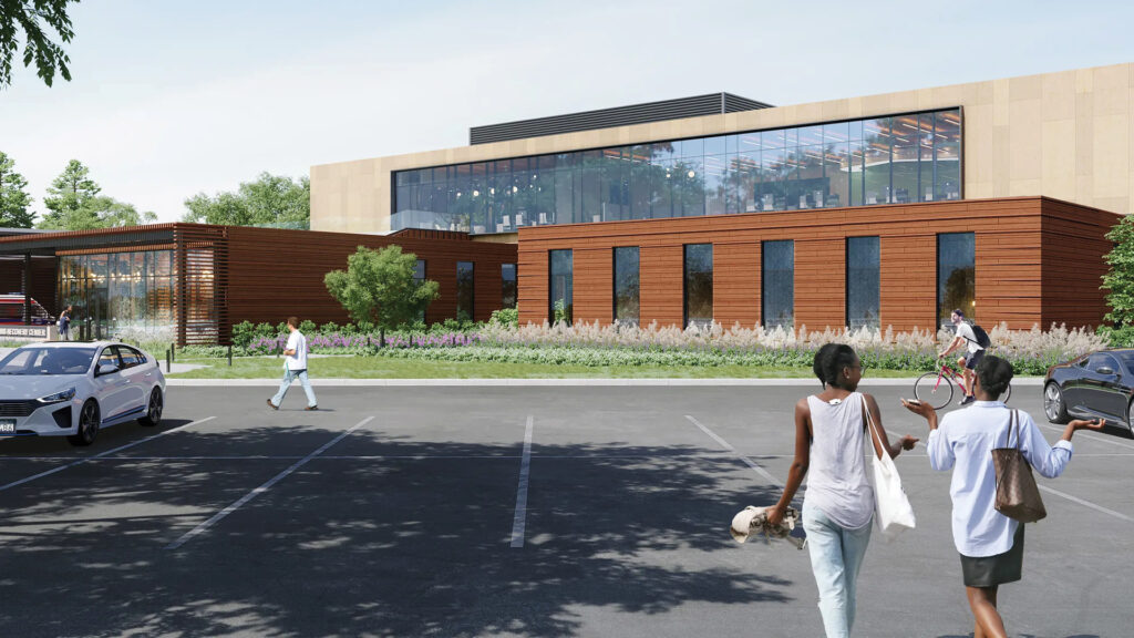 People walk and bike in a parking lot in front of a modern building with large windows and wooden paneling.