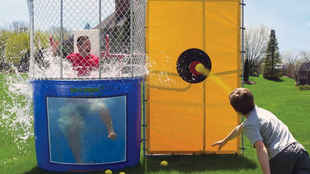 A boy throws a ball at a dunk tank target, splashing water as someone falls into the tank outdoors.