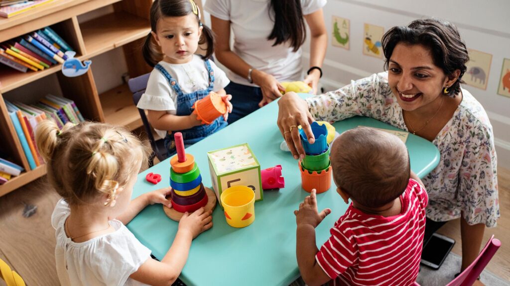 Smiling adult and four young children play with colorful toys at a small table in a classroom.