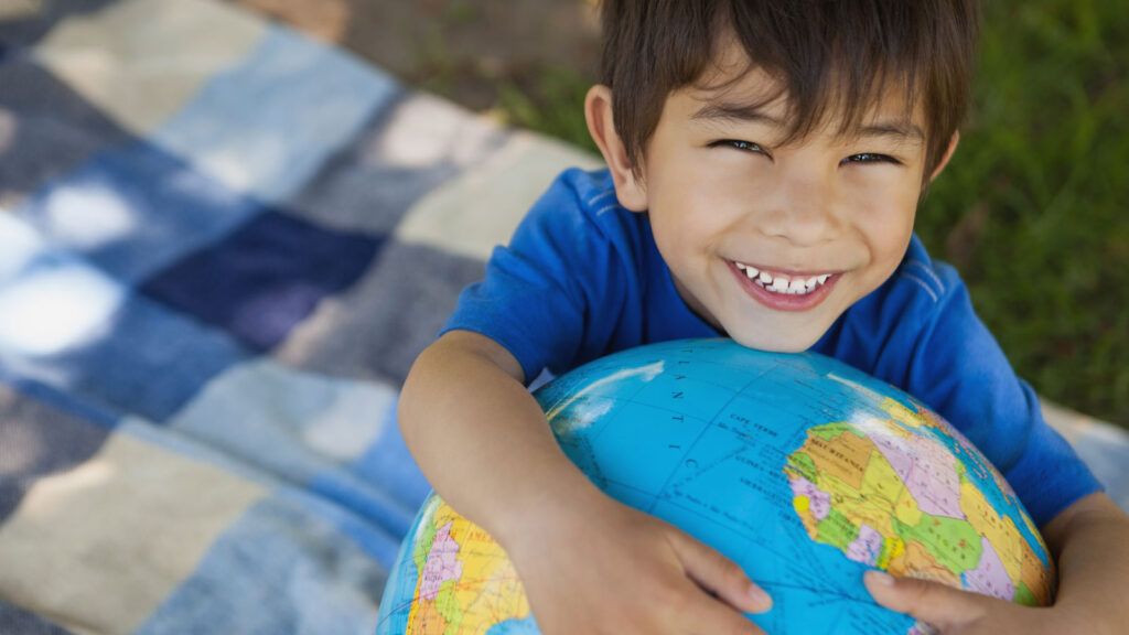 Smiling child in a blue shirt hugs a globe while sitting on a checkered blanket outdoors.