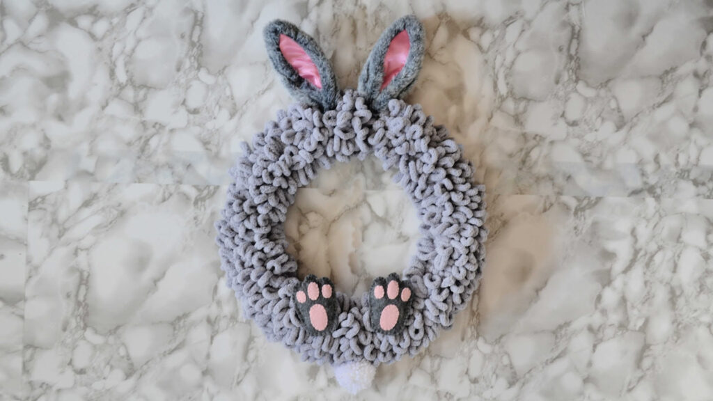 A fluffy gray wreath with bunny ears, pink paw prints, and a small white tail, on a marble surface.