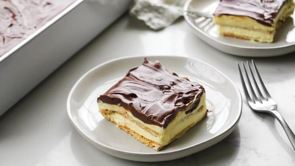 A slice of chocolate eclair cake with chocolate icing on a white plate with a fork beside it.