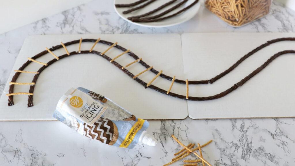 Licorice pieces and pretzel sticks arranged as a train track, with icing used to attach the pieces on a white surface.