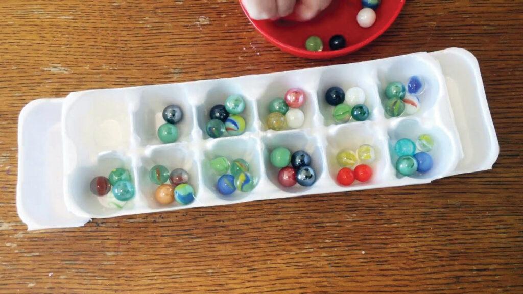 Ice cube tray filled with colorful marbles on a wooden table, with a red dish and hand above.