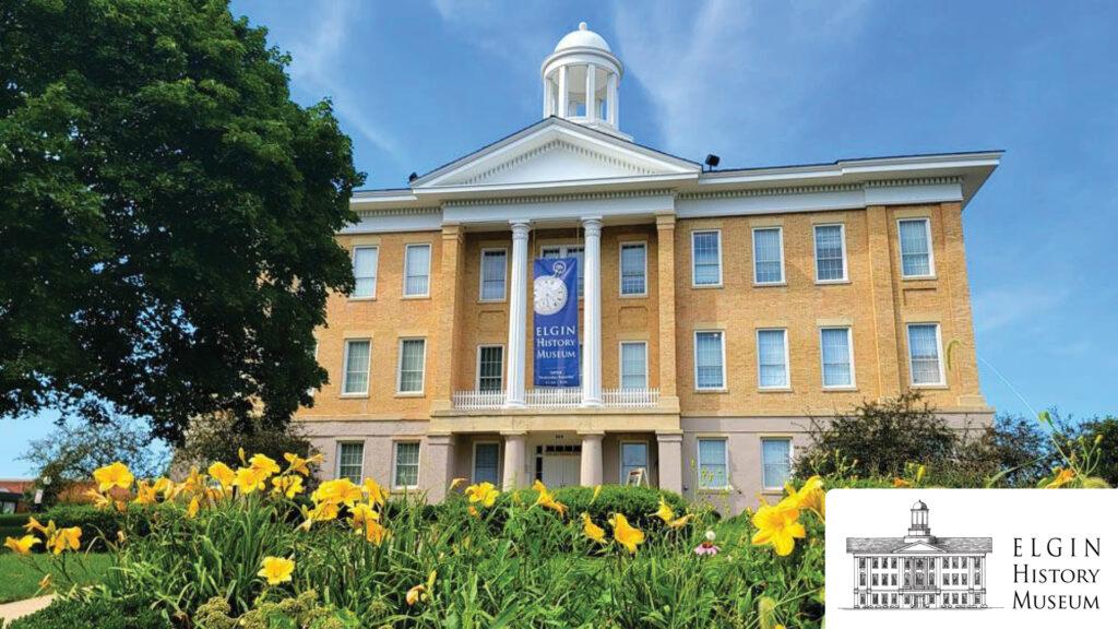 Yellow flowers bloom in front of the Elgin History Museum, a historic brick building with white columns and a dome.