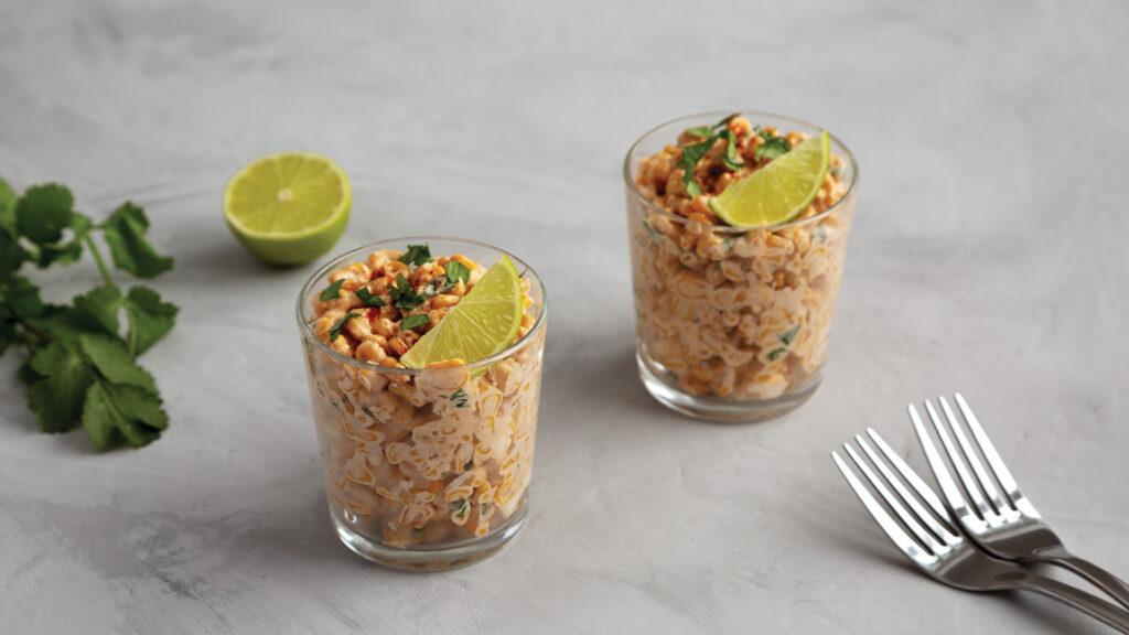 Two glasses filled with noodle salad, garnished with lime and herbs, with forks, cilantro, and lime nearby.