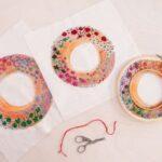 Three colorful embroidery hoops with floral patterns on fabric, accompanied by scissors and red thread on a table.