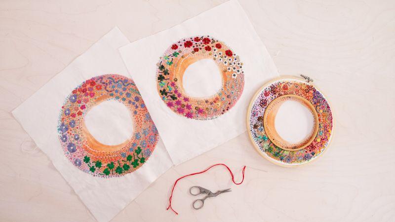 Three colorful embroidery hoops with floral patterns on fabric, accompanied by scissors and red thread on a table.