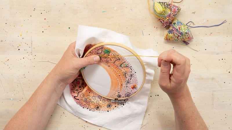 Hands embroidering colorful patterns on fabric in a hoop, with pincushions nearby.