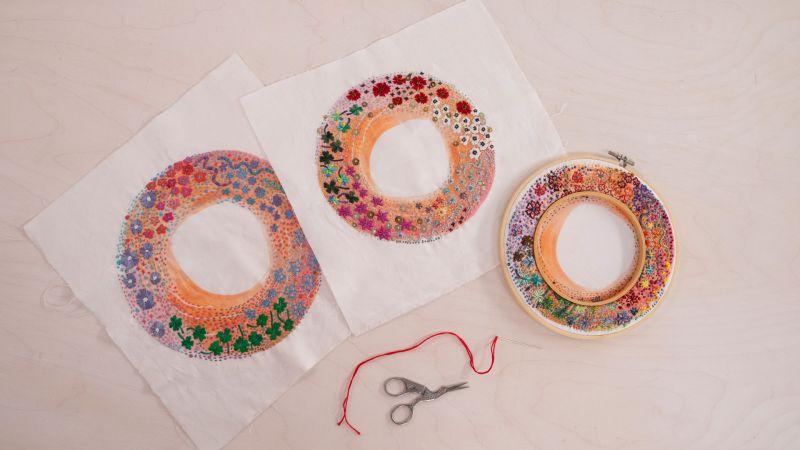 Two circular floral embroidery pieces on fabric, with a finished piece in a hoop, scissors, and red thread nearby.