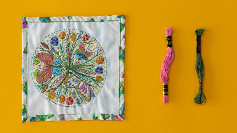 Colorful embroidery of butterflies and flowers on a square cloth, with pink and green embroidery threads beside it.