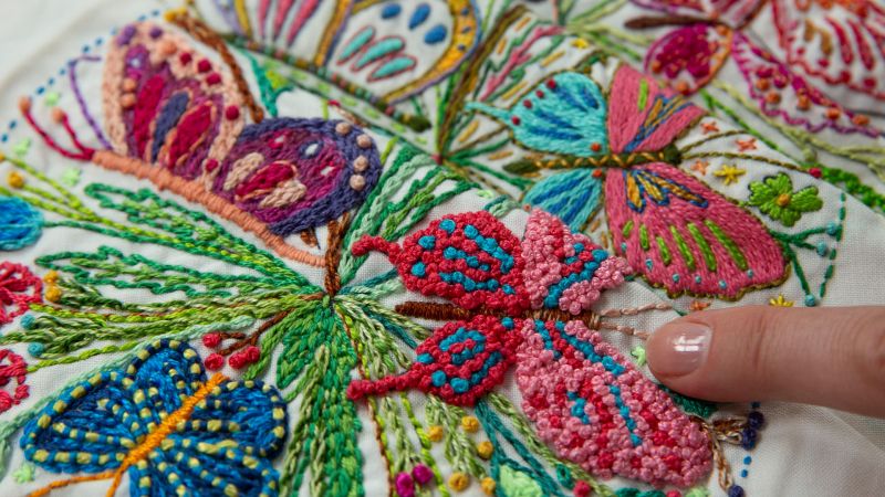 A finger points to colorful hand-embroidered butterflies on a fabric.