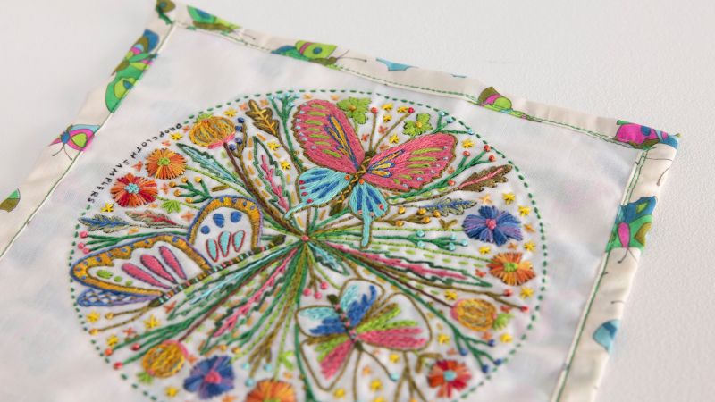 Colorful embroidery featuring butterflies, flowers, and greenery on a white square fabric with a patterned border.