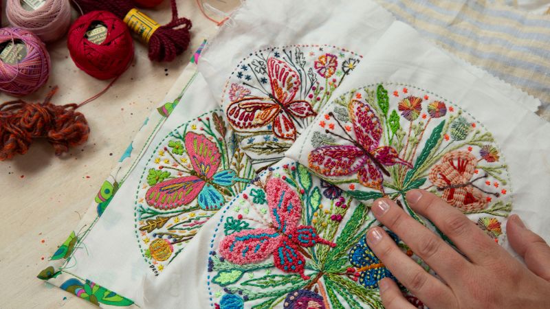 A hand touching a colorful embroidered butterfly design, surrounded by threads and yarn on a table.