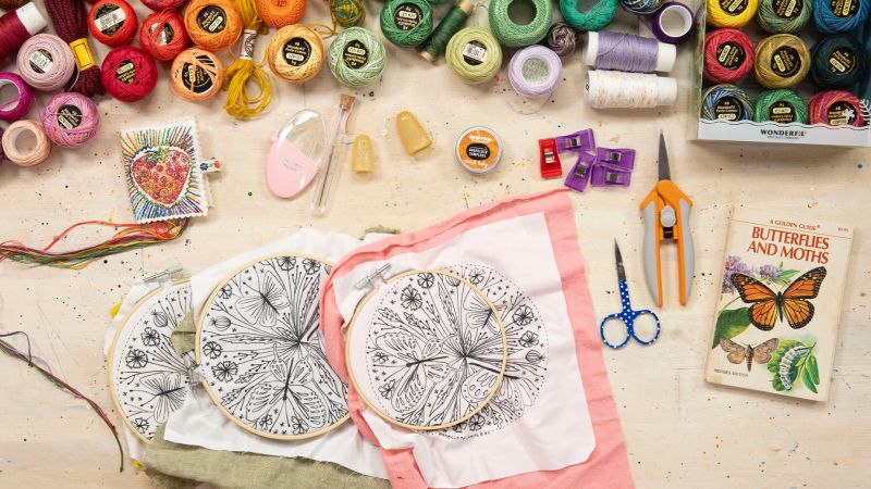 Embroidery supplies, threads, scissors, needles, hoops with patterns, and a “Butterflies and Moths” book on a table.