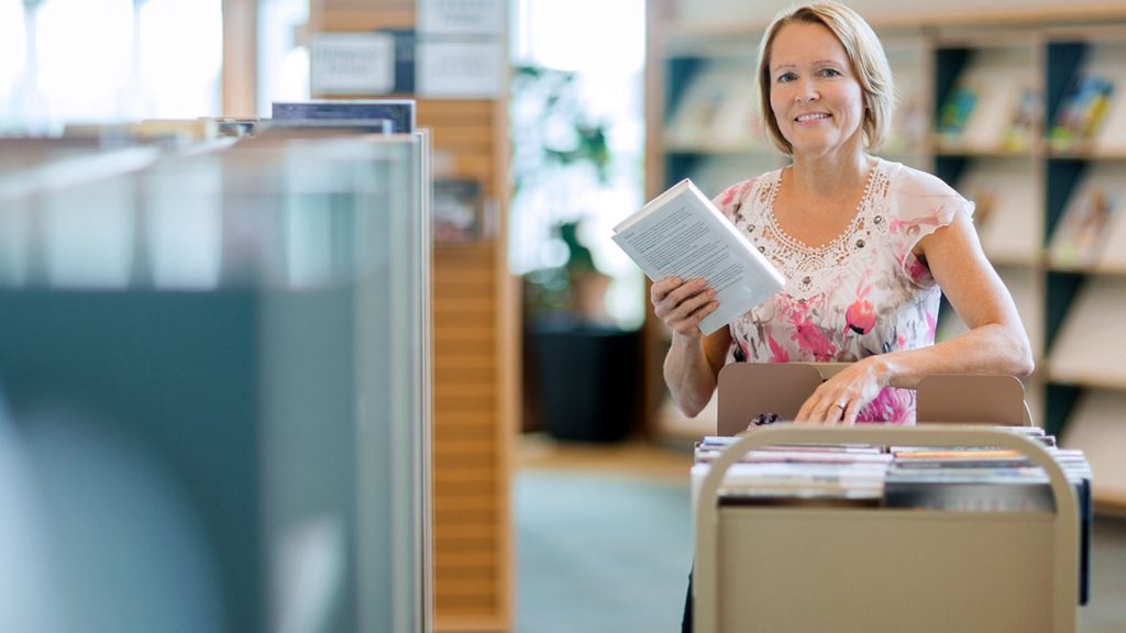 A woman is smiling while organizing books on a cart in a library.