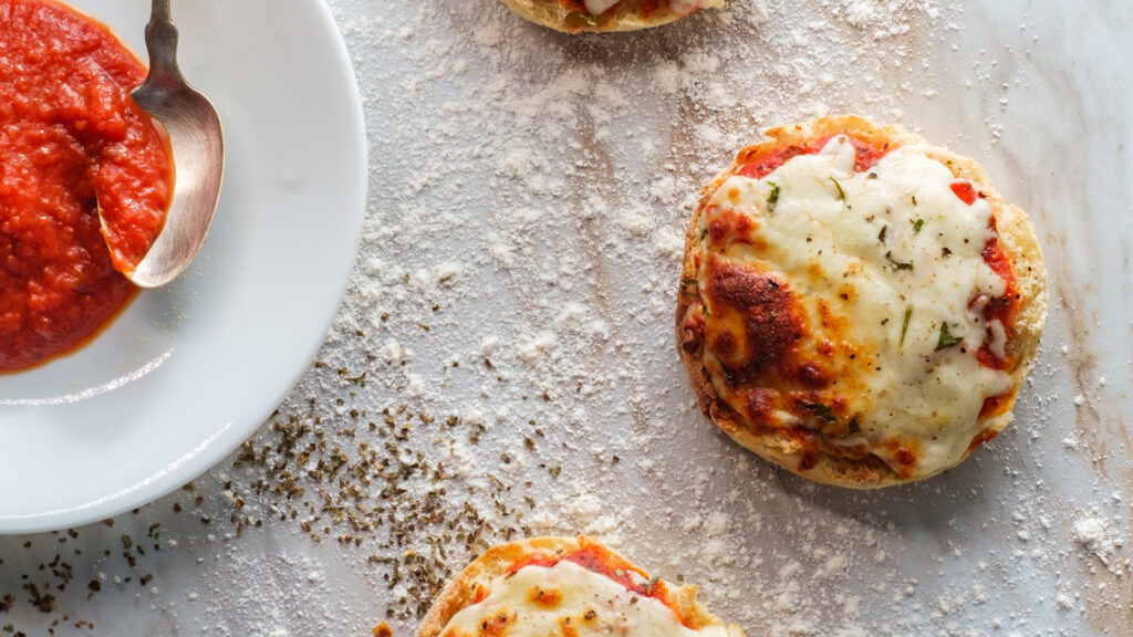 Mini pizzas with melted cheese on a floured surface, next to a plate of tomato sauce and a spoon.