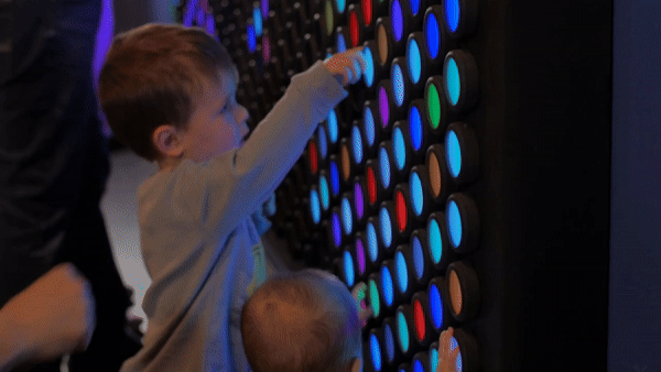 Two young children press colorful, glowing buttons on a large interactive wall panel.