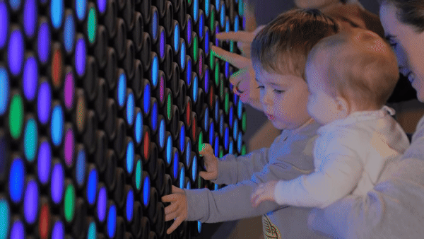 Two young children and an adult touch a wall of colorful, glowing lights at an interactive exhibit.