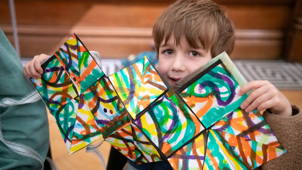 Young child holding up a colorful abstract painting made on folded paper, smiling indoors.