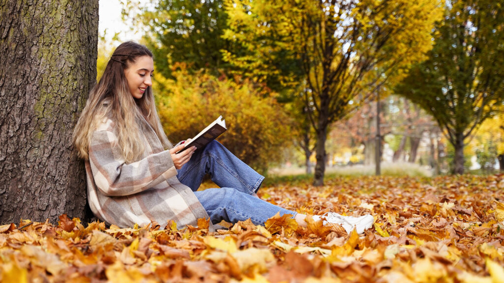 Woman sitting under a tree, reading a book amidst fallen autumn leaves, surrounded by trees with yellow foliage.