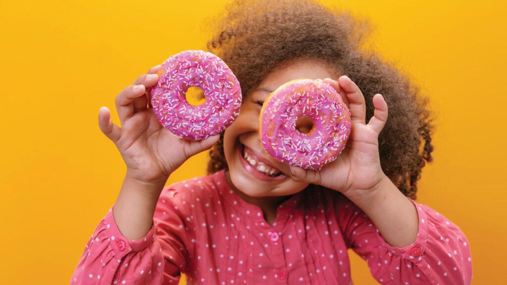 Smiling child holding two pink frosted donuts with sprinkles over their eyes against a yellow background.