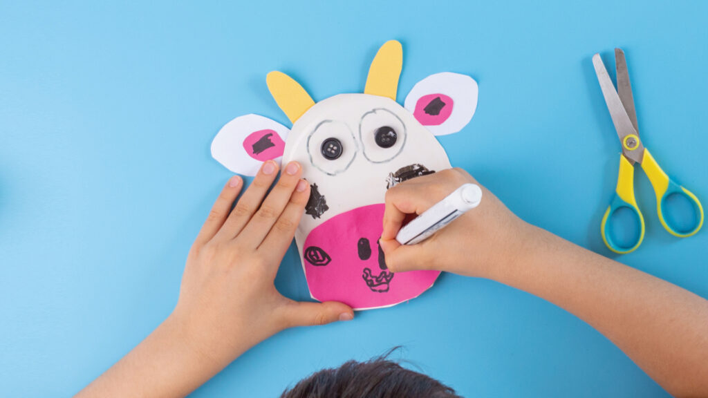 Child drawing a cow face on paper with marker, yellow scissors beside on a blue background.