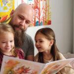 A man reads a colorful book to two young girls in a cozy room with bright artwork on the walls.