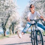 Woman joyfully cycling on a rural road lined with blossoming trees, wearing sunglasses and striped shirt.