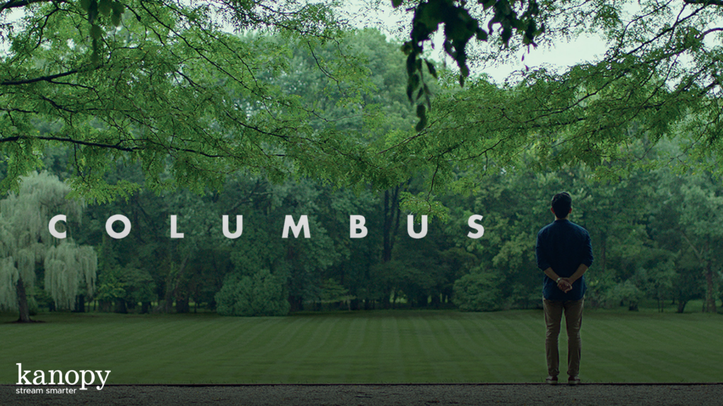 A person stands alone in a green park under trees, facing the word "COLUMBUS" in large letters.