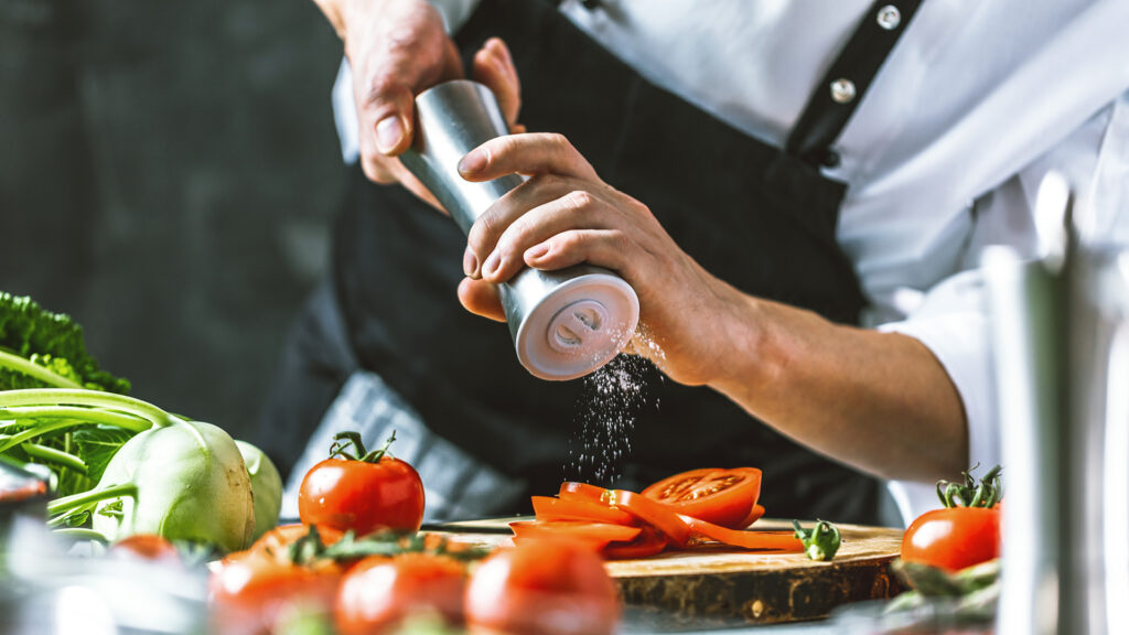 Chef sprinkling salt on sliced tomatoes on a wooden board, surrounded by fresh vegetables.