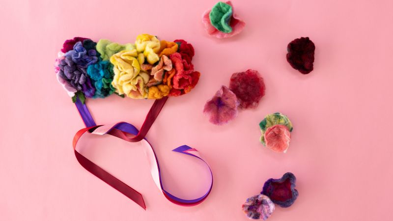 Colorful felt flowers on pink background with multicolored ribbons attached, and more flowers scattered around.