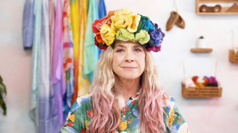 Person with long, wavy hair and a colorful floral headpiece, wearing a floral shirt, with rainbow fabrics in the background.