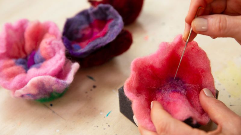 Close-up of hands using a needle to felt a pink and purple wool flower.