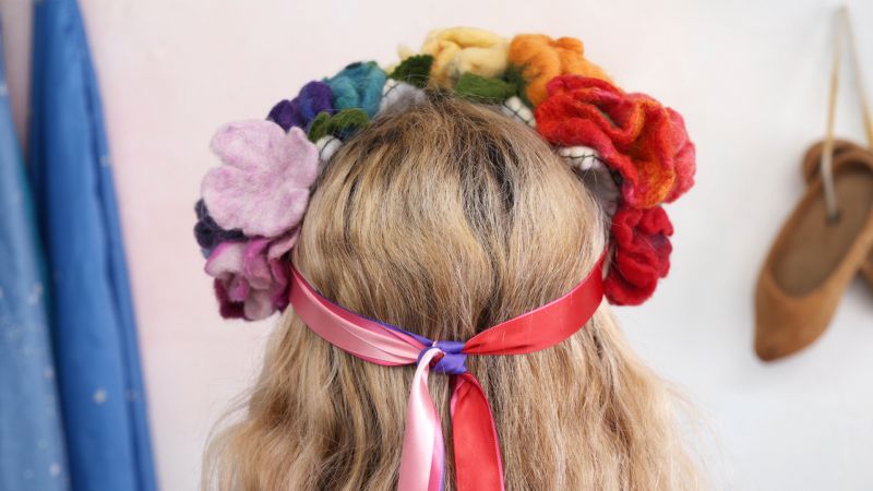 Blonde person with a colorful flower crown and red ribbons viewed from the back. A blue cloth and shoes hang in the background.