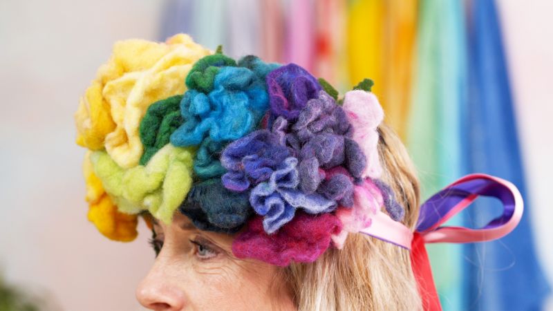 A person wearing a colorful flower crown made of felt, with vibrant ribbons hanging from it in the background.
